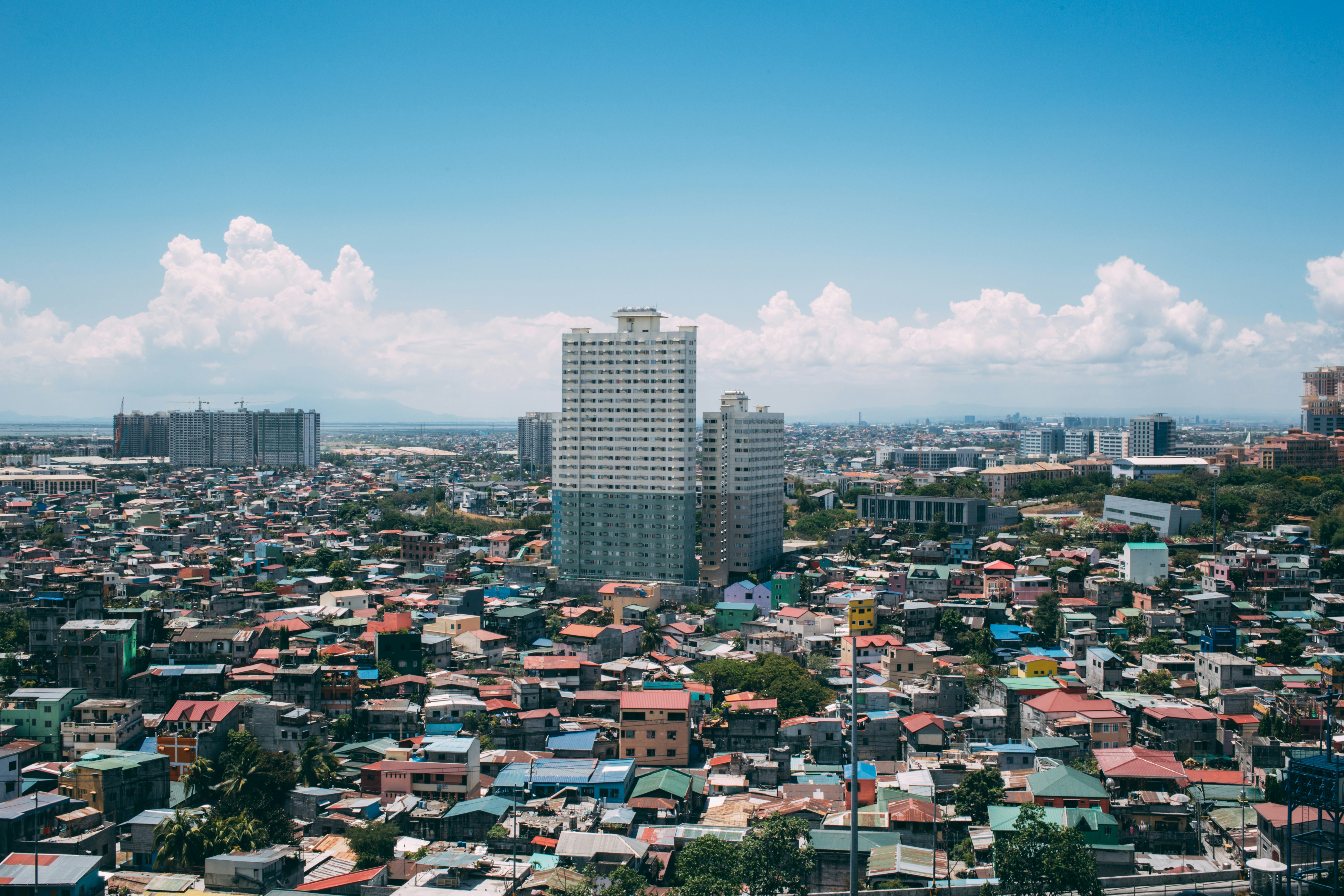 Aerial view of a densely populated urban neighborhood in Metro Manila, with a mix of low-rise residential houses and high-rise condominium buildings under a bright blue sky — representing the socioeconomic disparity and urban congestion addressed in Philippine reform proposals.