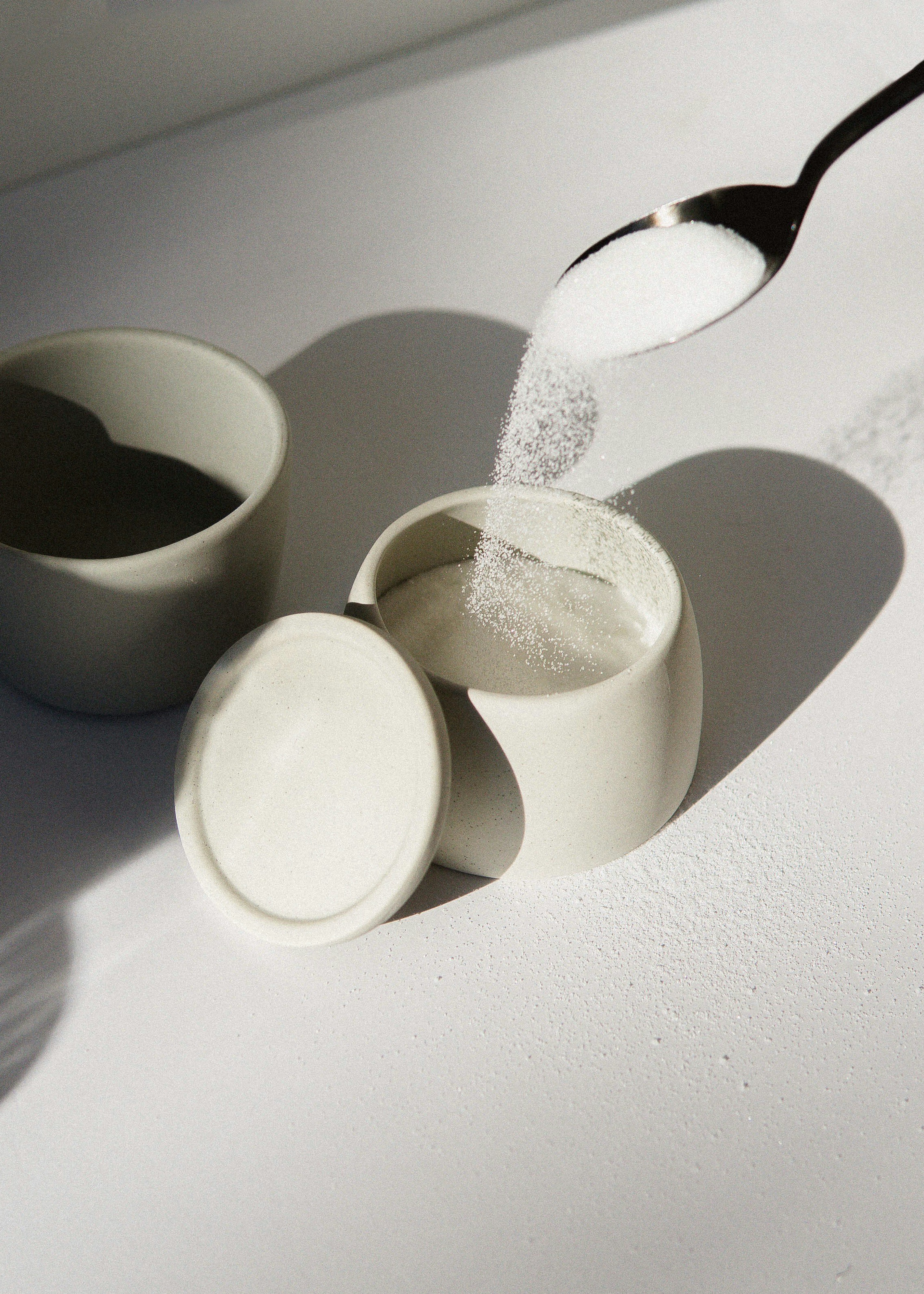 Granulated artificial sweetener being poured from a spoon into a ceramic container, representing the widespread dietary presence of substances like aspartame and their potential link to mental health disorders such as ADHD and OCD.