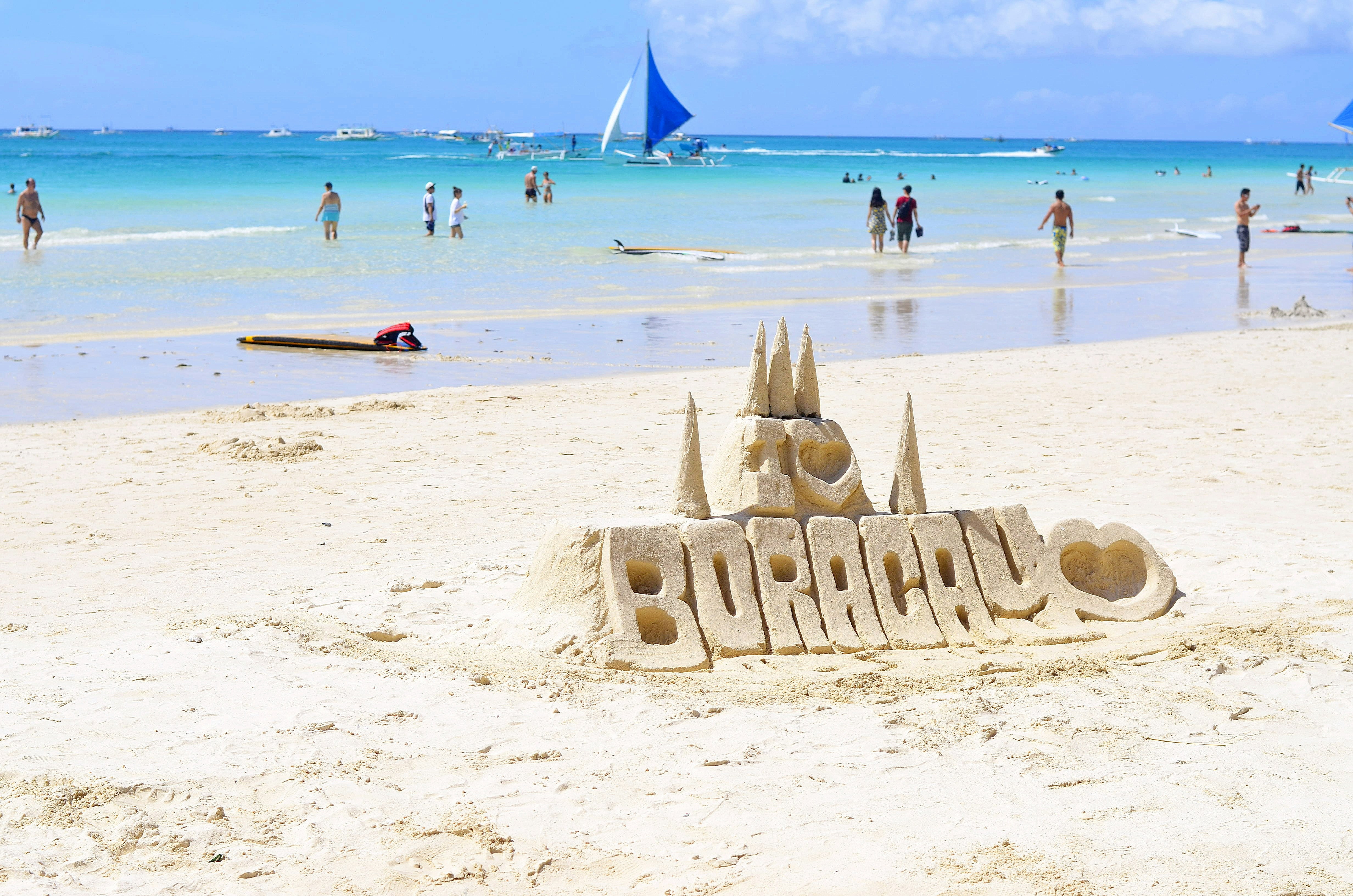 Sandcastle sculpture reading “I ❤️ Boracay” on White Beach, with people swimming and sailboats on turquoise water in the background — a classic scene from Boracay Island, Aklan.