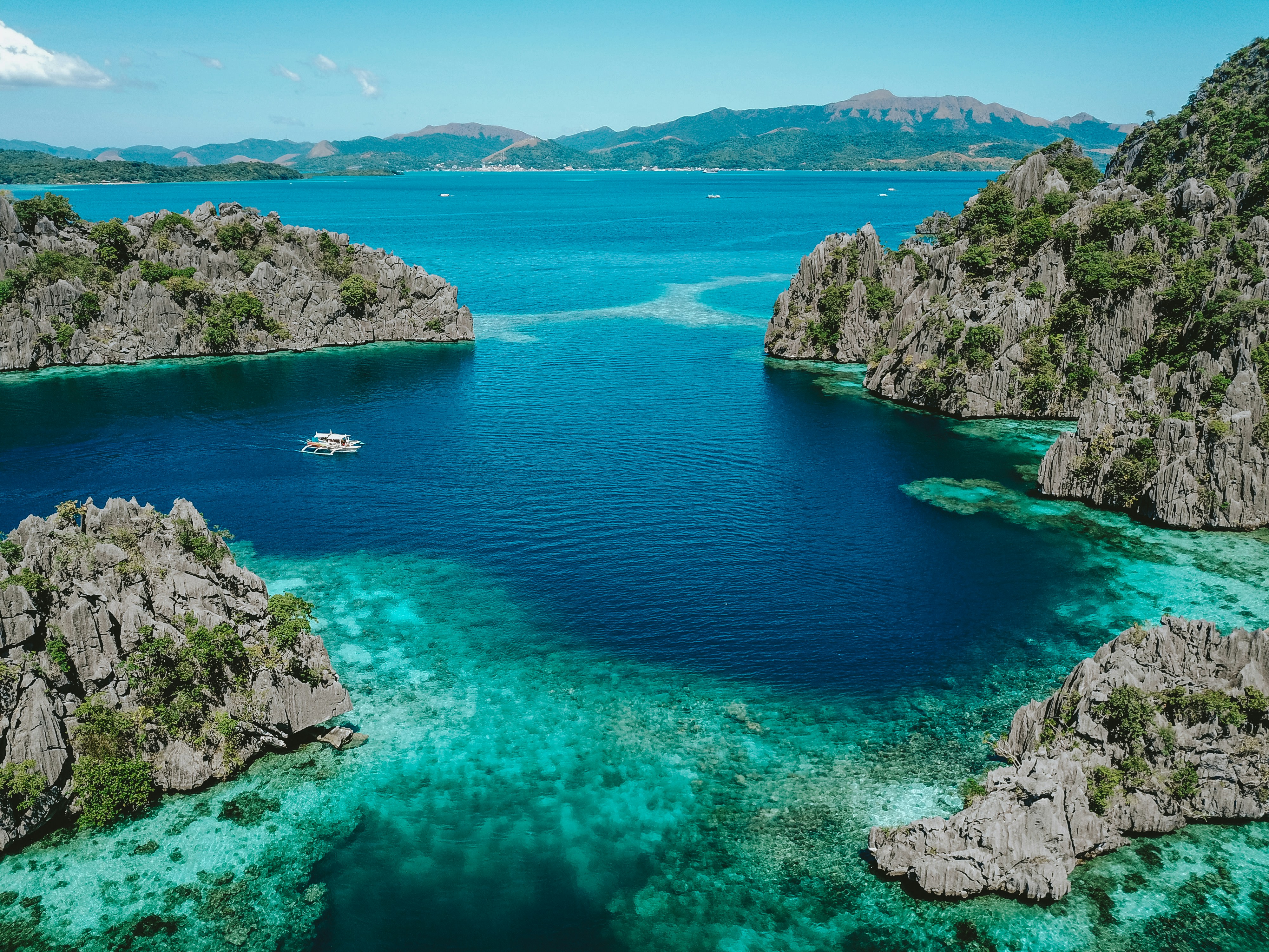 Aerial view of Coron Bay in Palawan, featuring limestone cliffs, turquoise reefs, and a traditional Filipino outrigger boat sailing through the clear blue water.