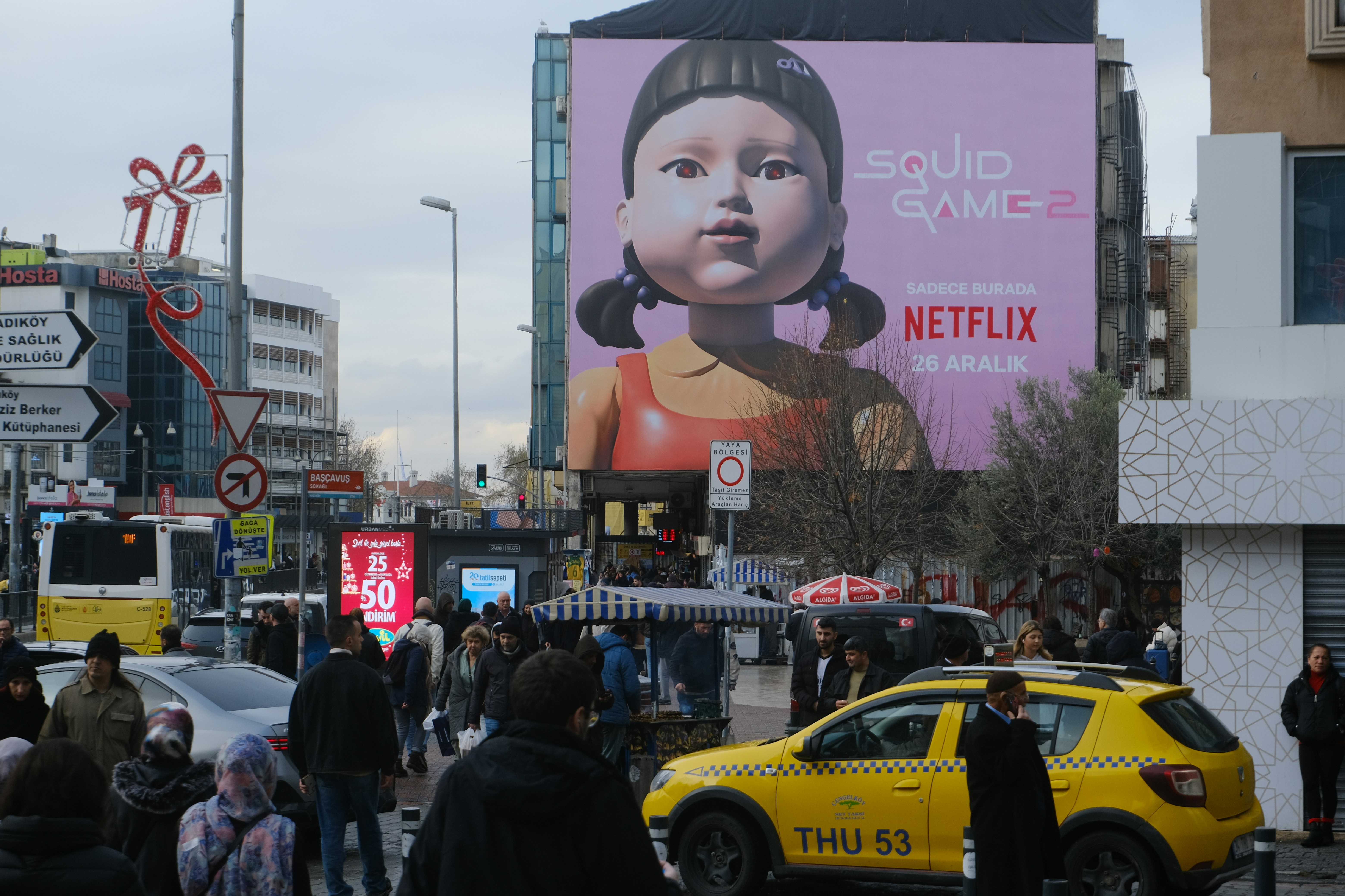 Crowded urban street scene featuring a massive Netflix billboard of the Squid Game robot doll, illustrating the global cultural reach of Korean media and its presence in everyday public life.