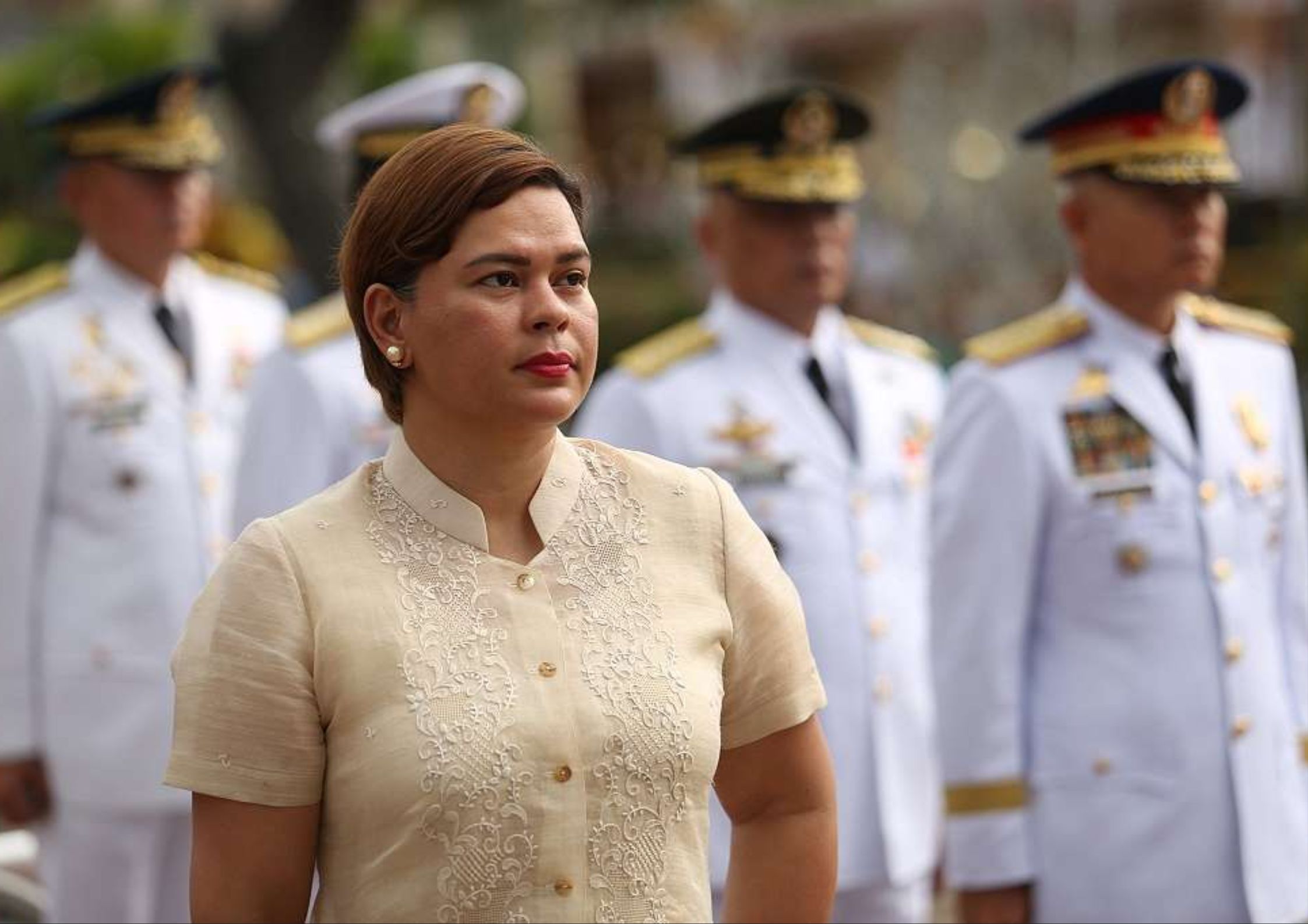 Philippine Vice President Sara Duterte in a formal barong-style blouse standing in front of uniformed military officials, symbolizing her embattled but resilient position amid impeachment proceedings and political tensions.