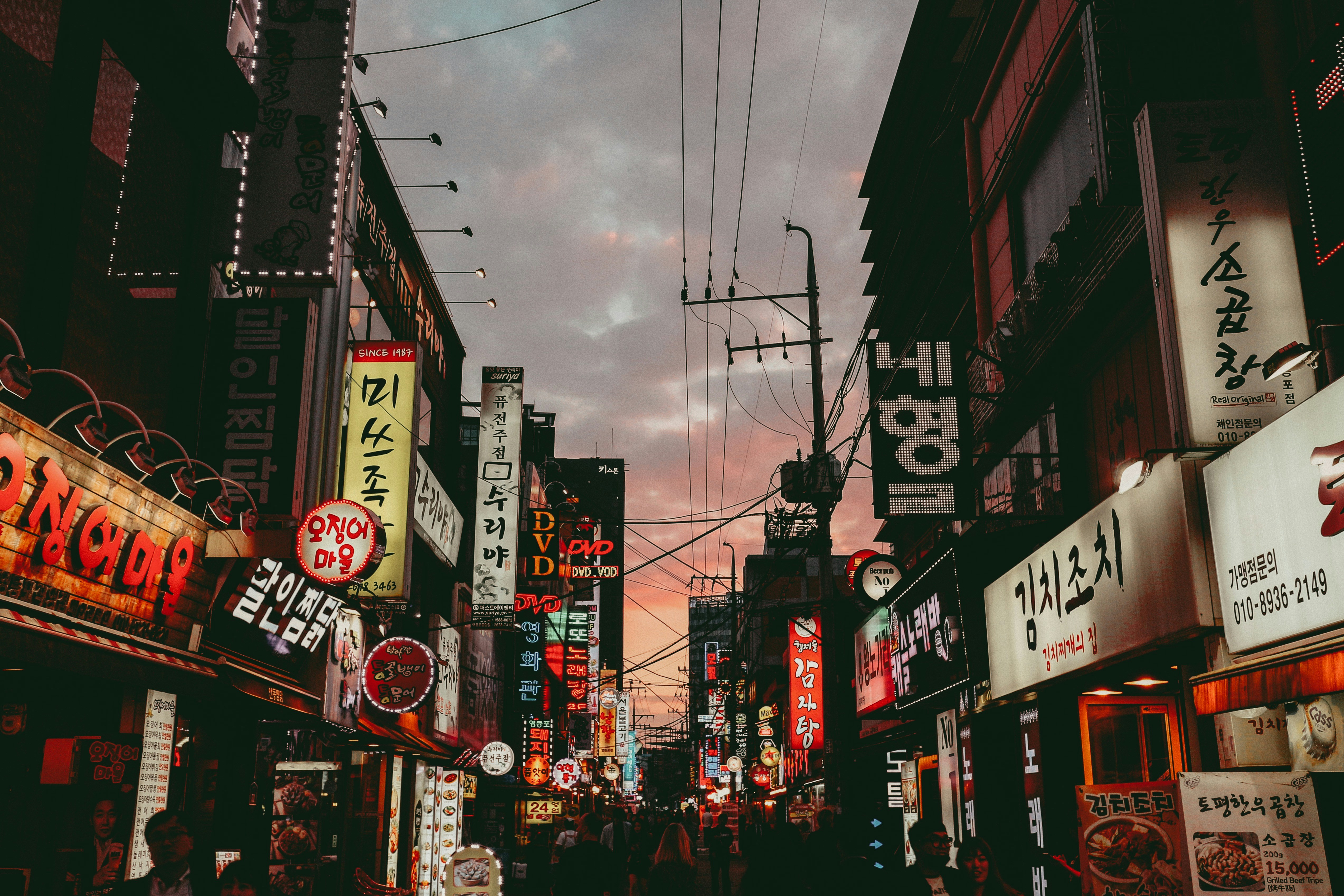 A densely lit urban street in South Korea at dusk, glowing with neon signage in Hangul. Restaurants, karaoke bars, and adult entertainment venues line both sides of the alley as electrical wires crisscross the sky—symbolizing the commercial vibrance and nightlife culture that has extended its reach into Southeast Asian cities like Angeles, Subic, and Cebu.