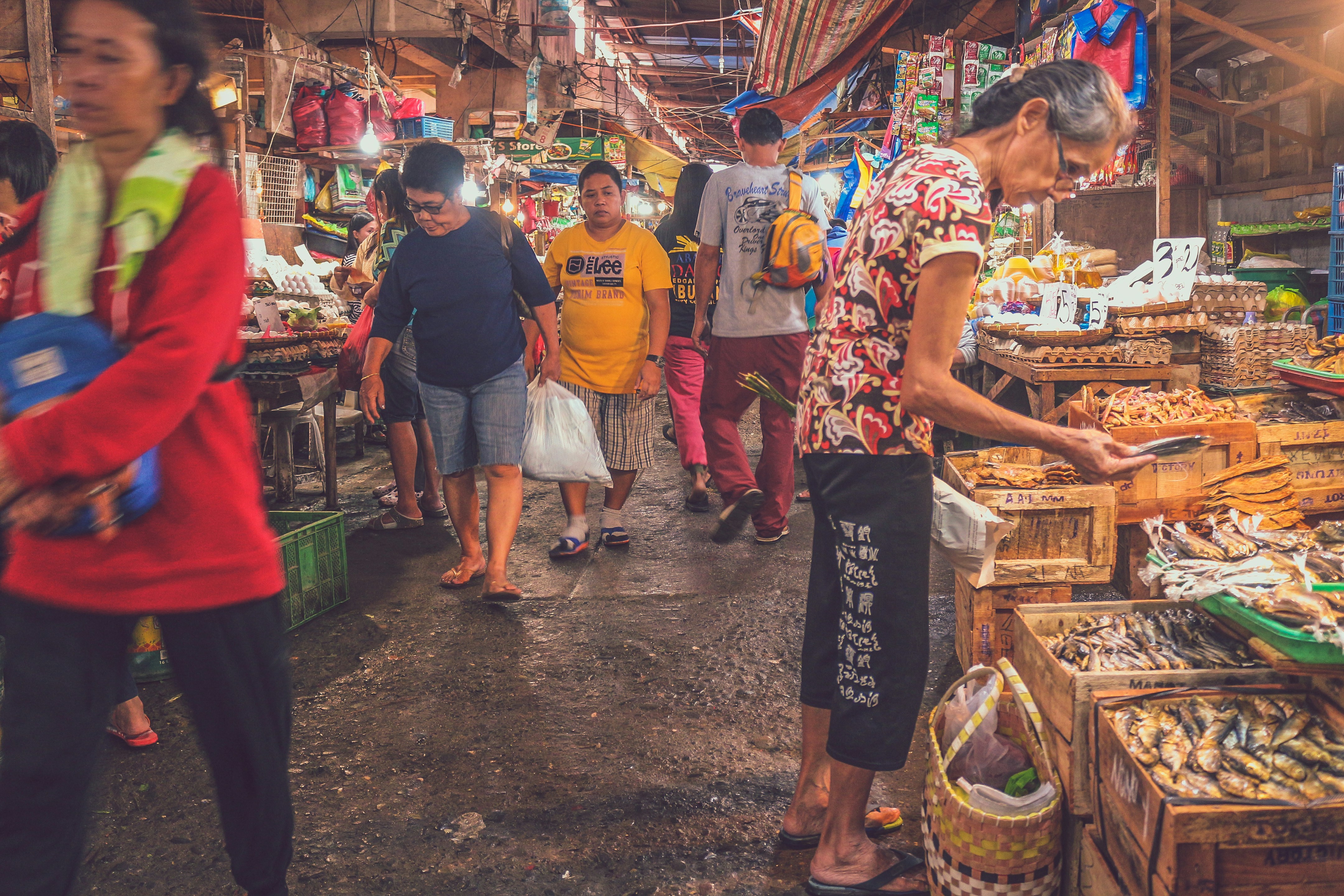 Filipino wet market scene bustling with activity — an elderly woman selects dried fish from a wooden crate, while shoppers in casual clothes navigate narrow aisles filled with local produce, eggs, and other goods under a canopy of makeshift tarps and hanging lights.